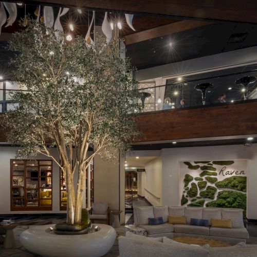 An upscale indoor lobby with a large planted tree, warm lights, wooden balconies, and a &ldquo;Raven&rdquo; greenery wall mural.