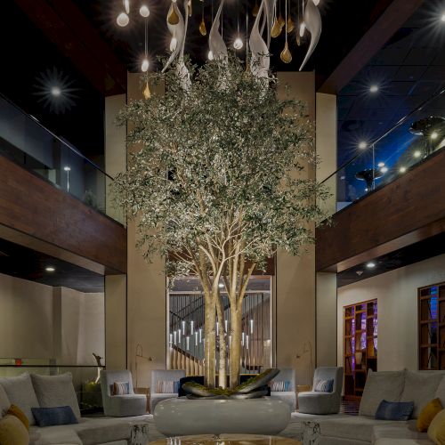 An upscale lobby atrium with a tall indoor olive tree, hanging lights, modern stairs/railings, and sofas under a warm glow.