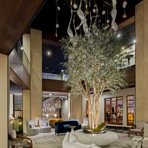 An elegant indoor atrium lobby with a lit chandelier, hanging spoons, olive tree, and modern seating under skylights.