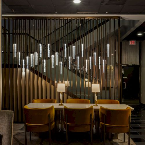A modern hotel lounge with a long table, yellow chairs, and a hallway of glowing vertical glass slats with an EXIT sign.