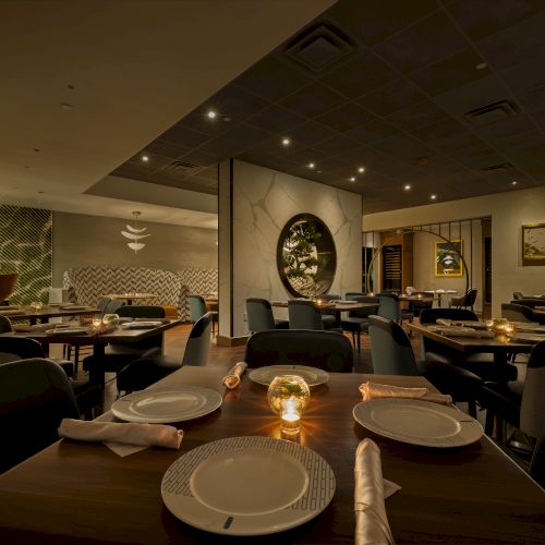 An elegant restaurant dining room with neatly set tables, dark chairs, warm ceiling lights, and framed wall art.