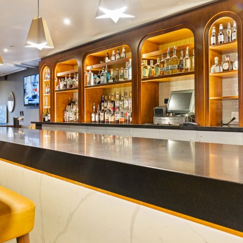 A modern hotel bar with illuminated wooden shelves stocked with liquor bottles, plus a countertop sink and taps.