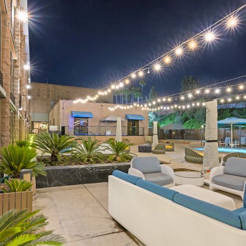 Nighttime hotel courtyard with string lights, lounge seating, plants, and a glowing pool area.