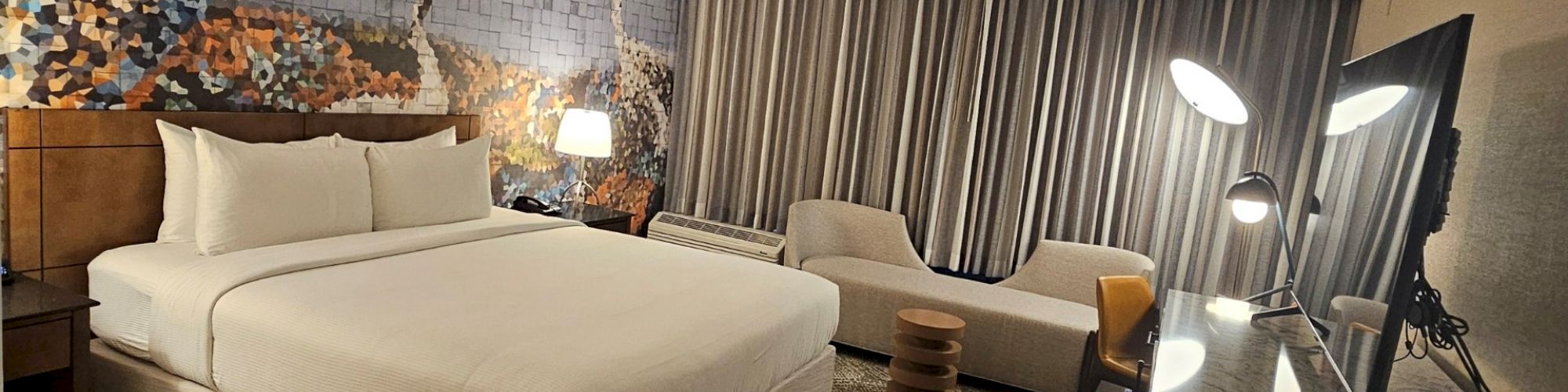 A hotel room with a bed, decorative wall art, a seating area, and a TV. Modern decor with a patterned carpet and stylish lighting.