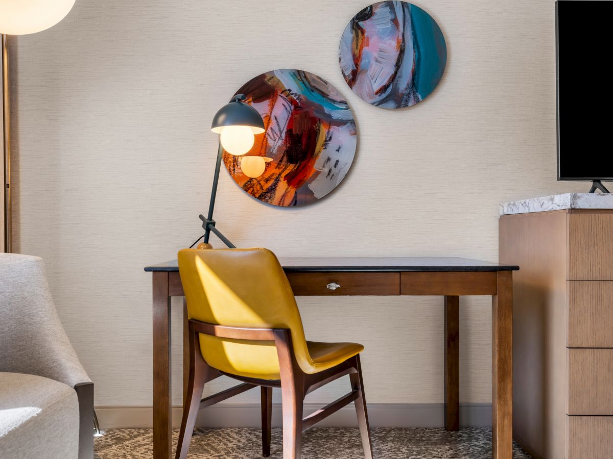 A modern room with a wooden desk, yellow chair, desk lamp, abstract wall art, and a TV on a dresser.