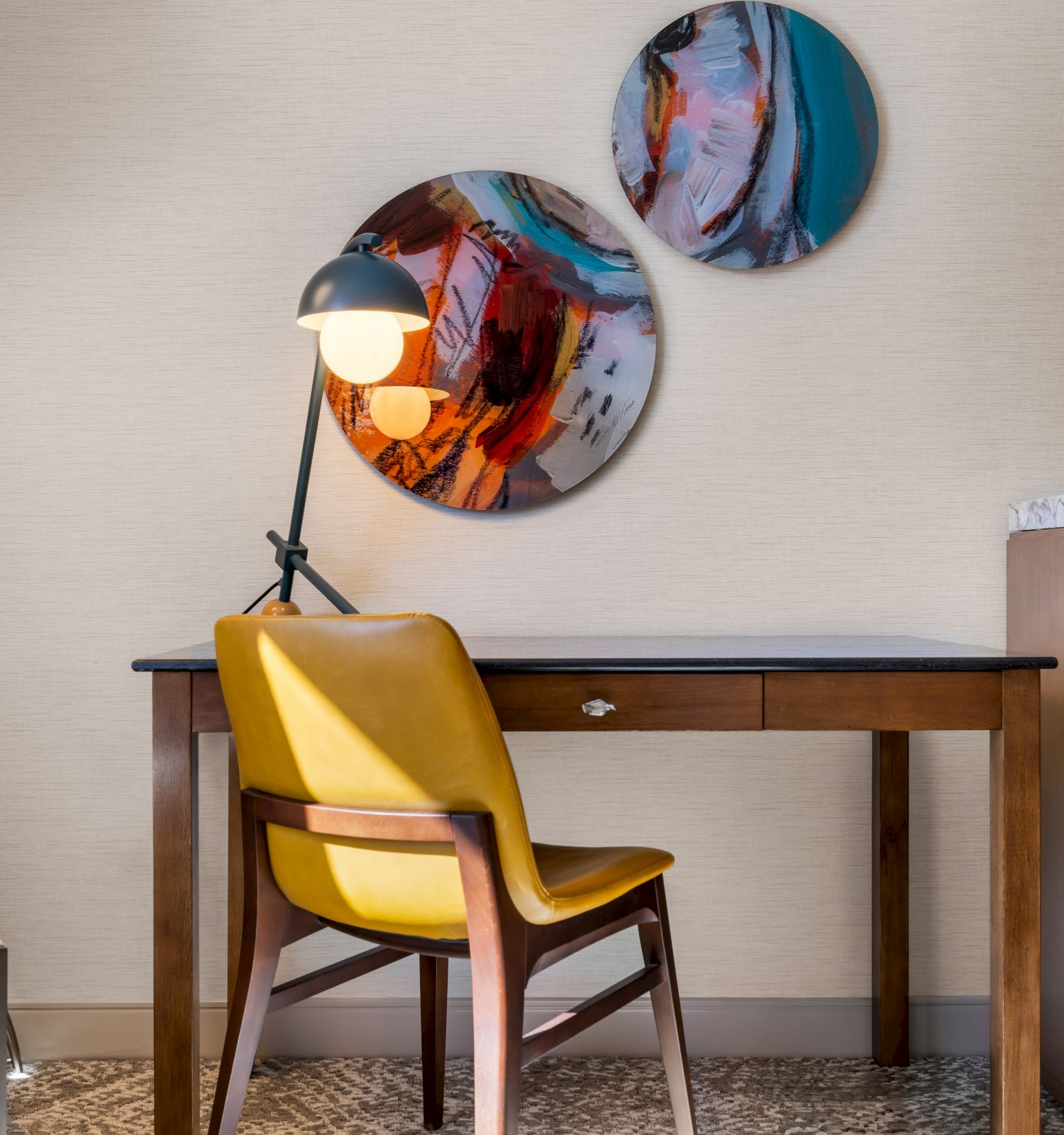 A modern workspace with a wooden desk, yellow chair, lamp, abstract art on the wall, and a TV atop a cabinet.