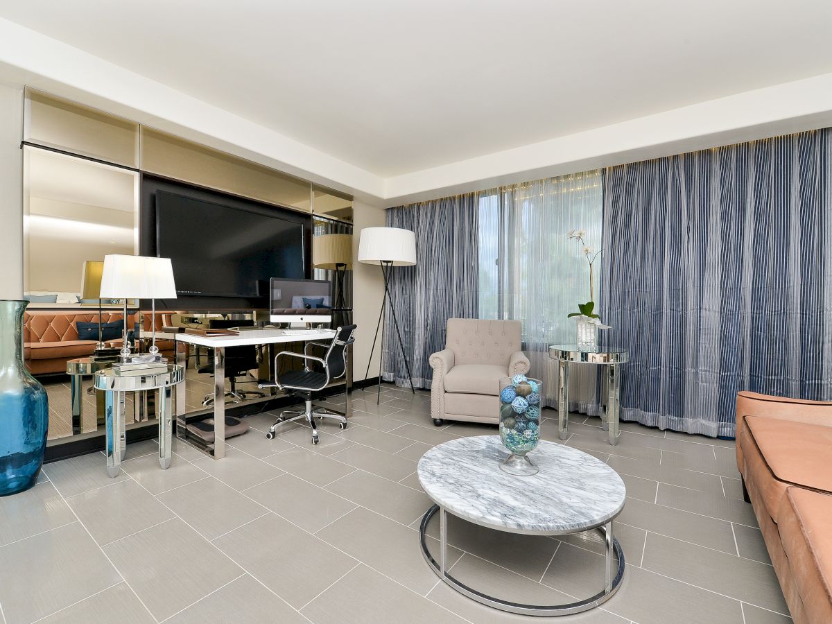 A living space with a grey armchair, large TV, desk, floor lamps, and a round marble coffee table. Blue drapes cover a large window.