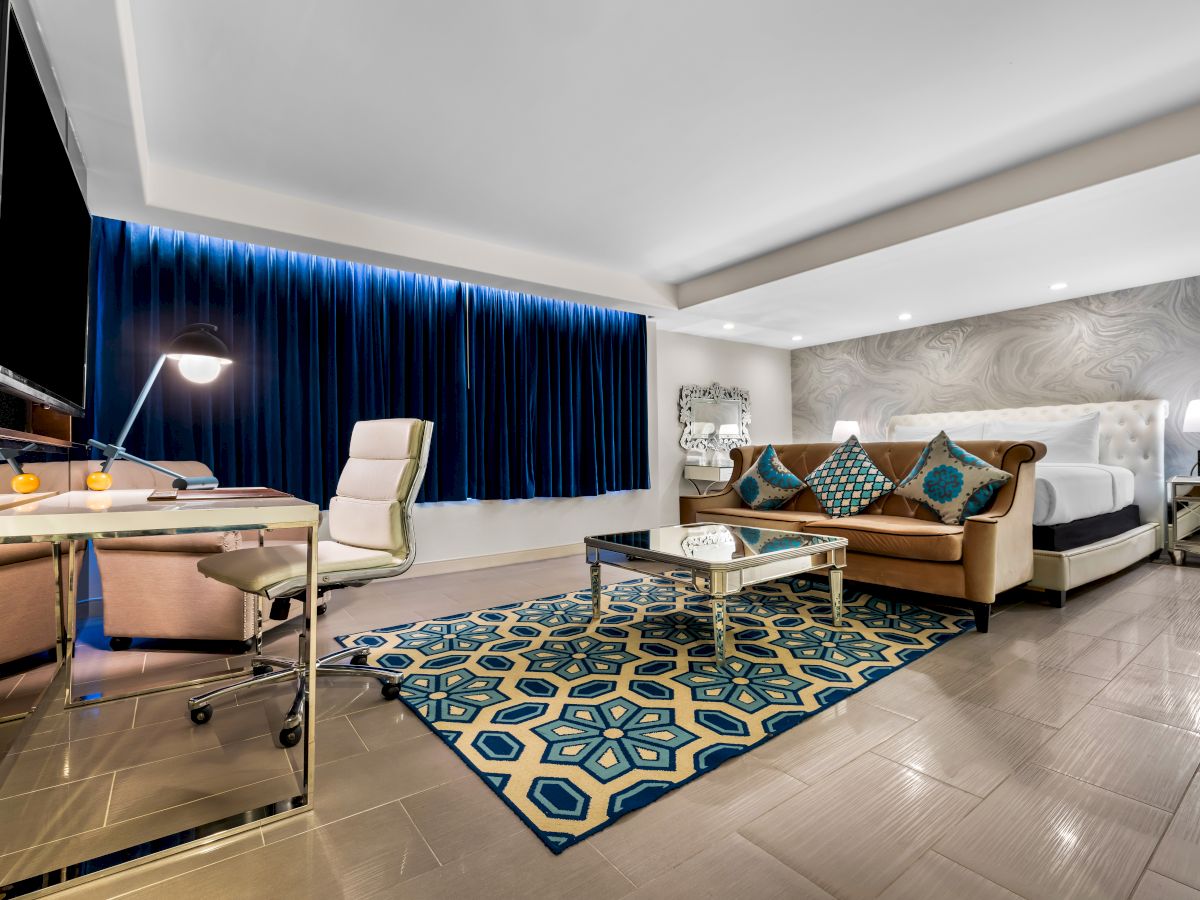 A modern hotel room with a bed, a sofa, a desk, a lamp, and a TV. The decor features blue curtains and a patterned rug.