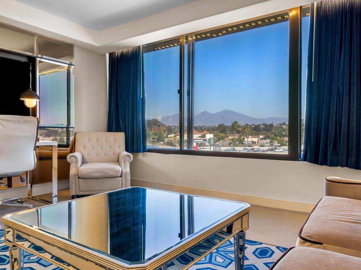 A modern room with a beige armchair, mirrored table, and a large window offering a city and mountain view.