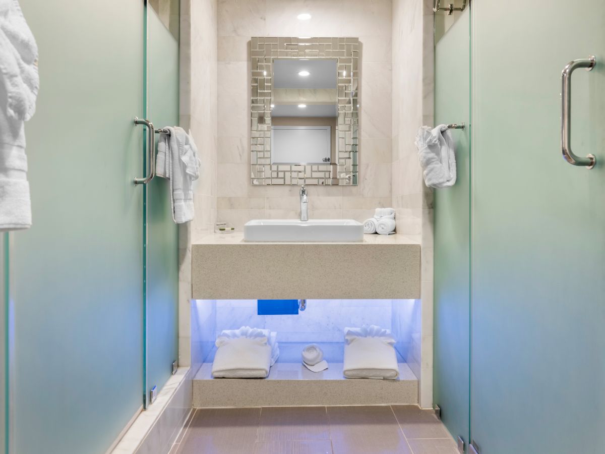 A modern bathroom with frosted glass doors, a sink and mirror, and neatly folded towels under the vanity with soft lighting.