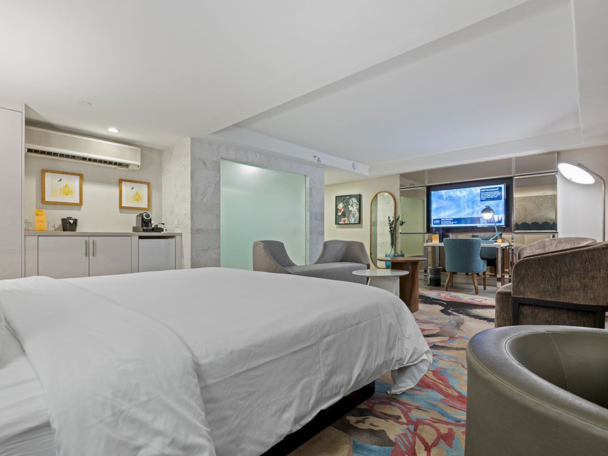A modern hotel room with a king bed, kitchenette, seating area, and a flat-screen TV.