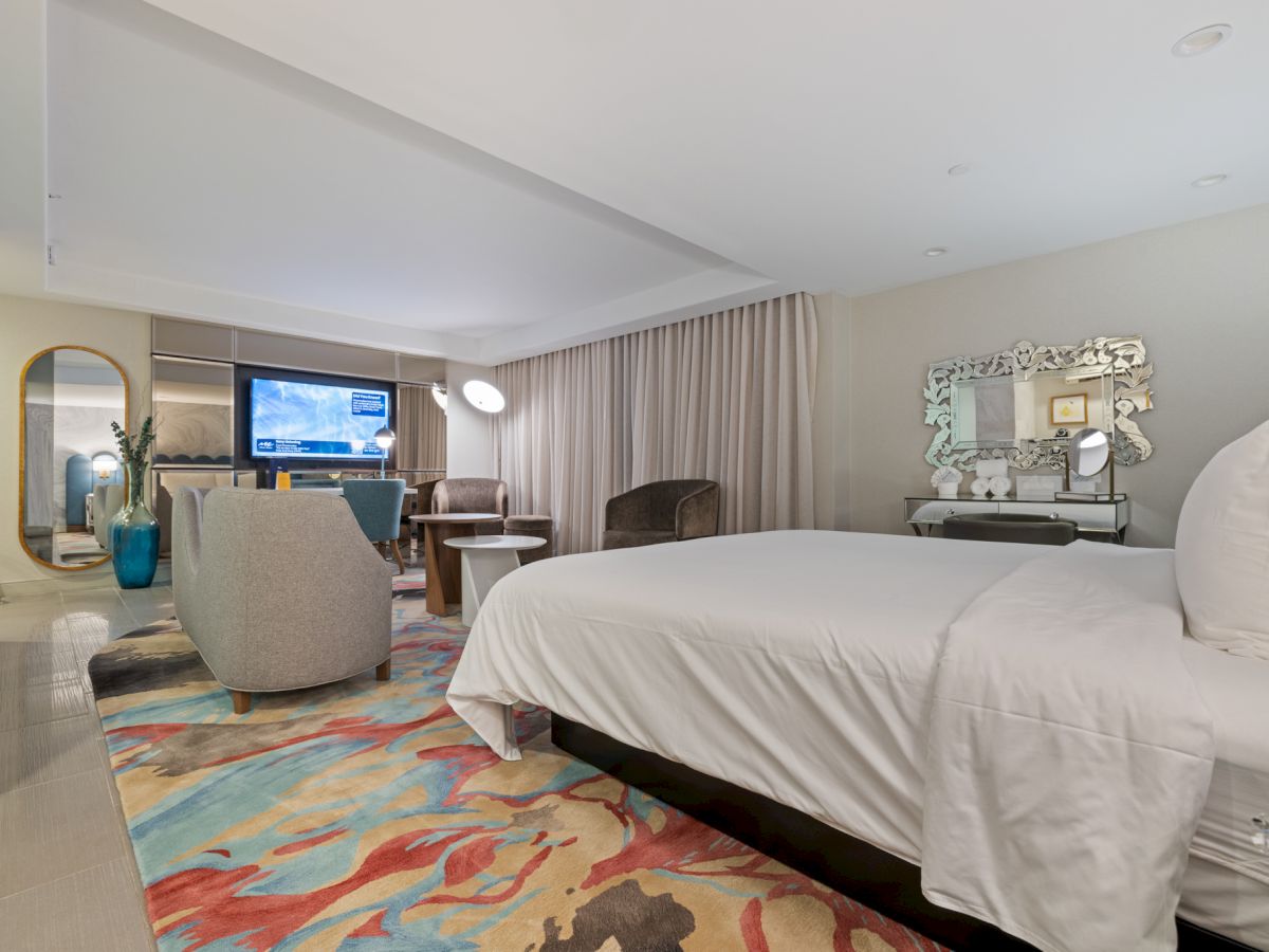 A modern hotel suite with a king bed, vanity mirror, TV lounge area, and soft curtains under recessed lights.