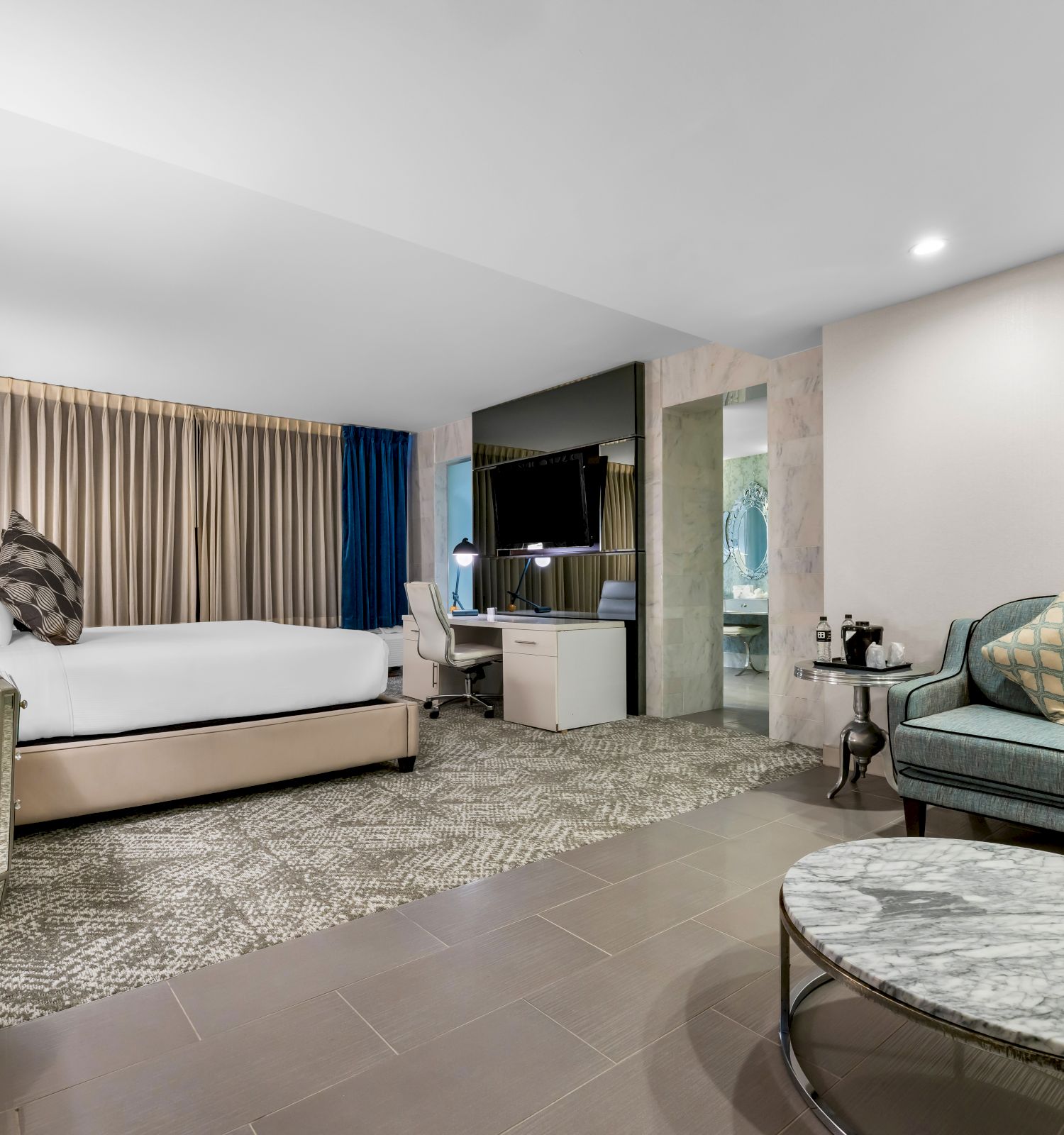 A modern hotel room with a bed, sofa, coffee table, desk, and TV, featuring neutral tones and stylish decor, all well-lit.