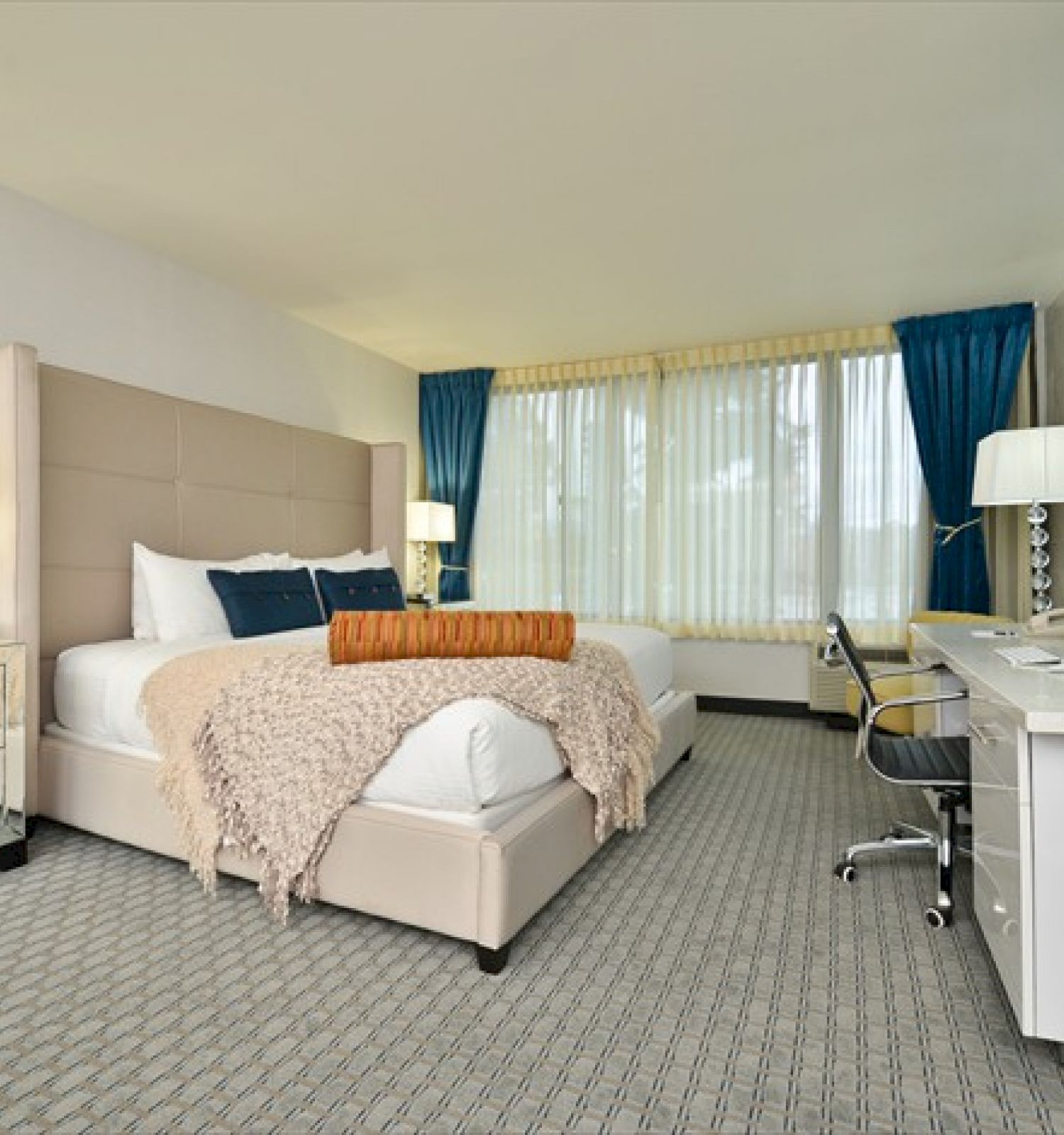 A modern hotel room with a large bed, desk, chair, lamps, and windows with blue curtains. End of sentence.