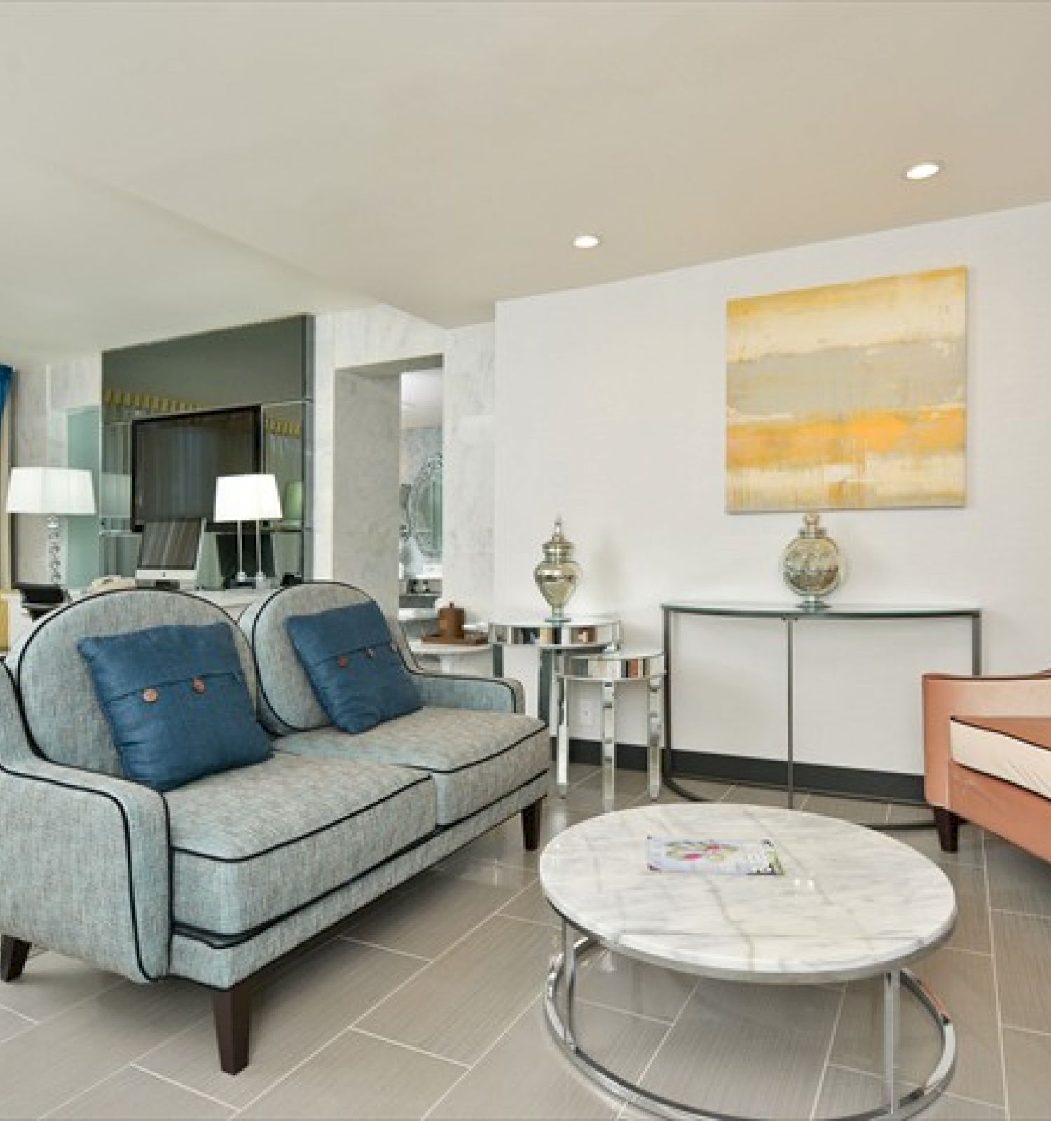 A living room with a sofa, armchair, coffee table, console table, lamps, and wall art. There is also a bed in the background.