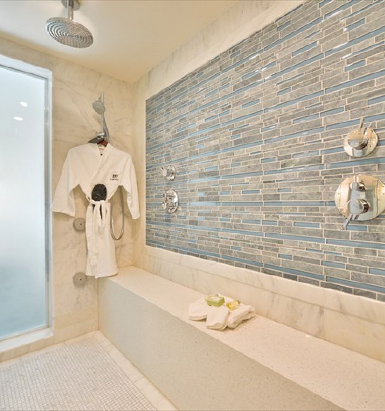 A luxurious shower room with modern fixtures, a bench, a robe hanging, and folded towels on the bench, with a frosted glass door.