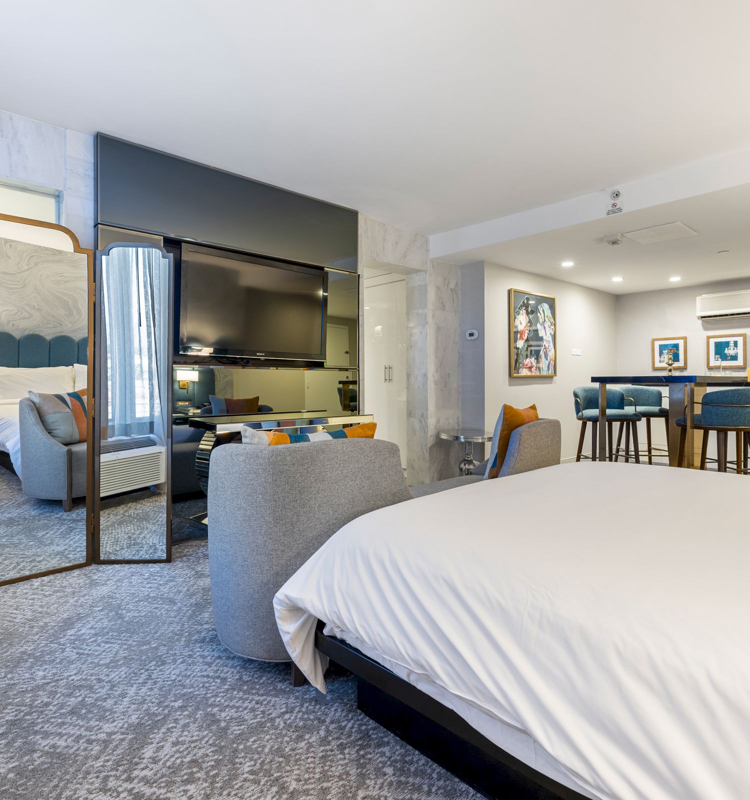 Modern hotel suite with a bed, flat-screen TV, lounge seating, bar counter, and blue chairs beside marble walls and decor.