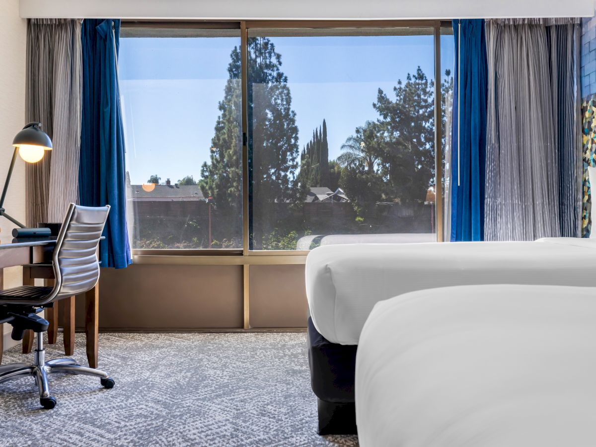 The image shows a hotel room with two beds, a desk with a lamp, and a large window with a view of trees outside.