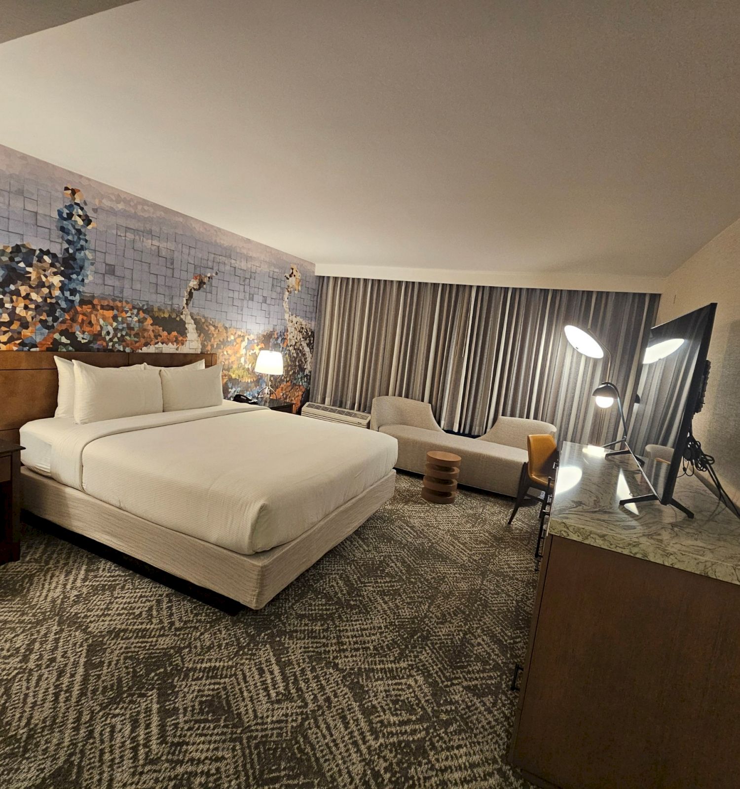 A modern hotel room with a bed, artwork on the wall, a sofa, a desk, and a TV, featuring stylish decor and lighting.