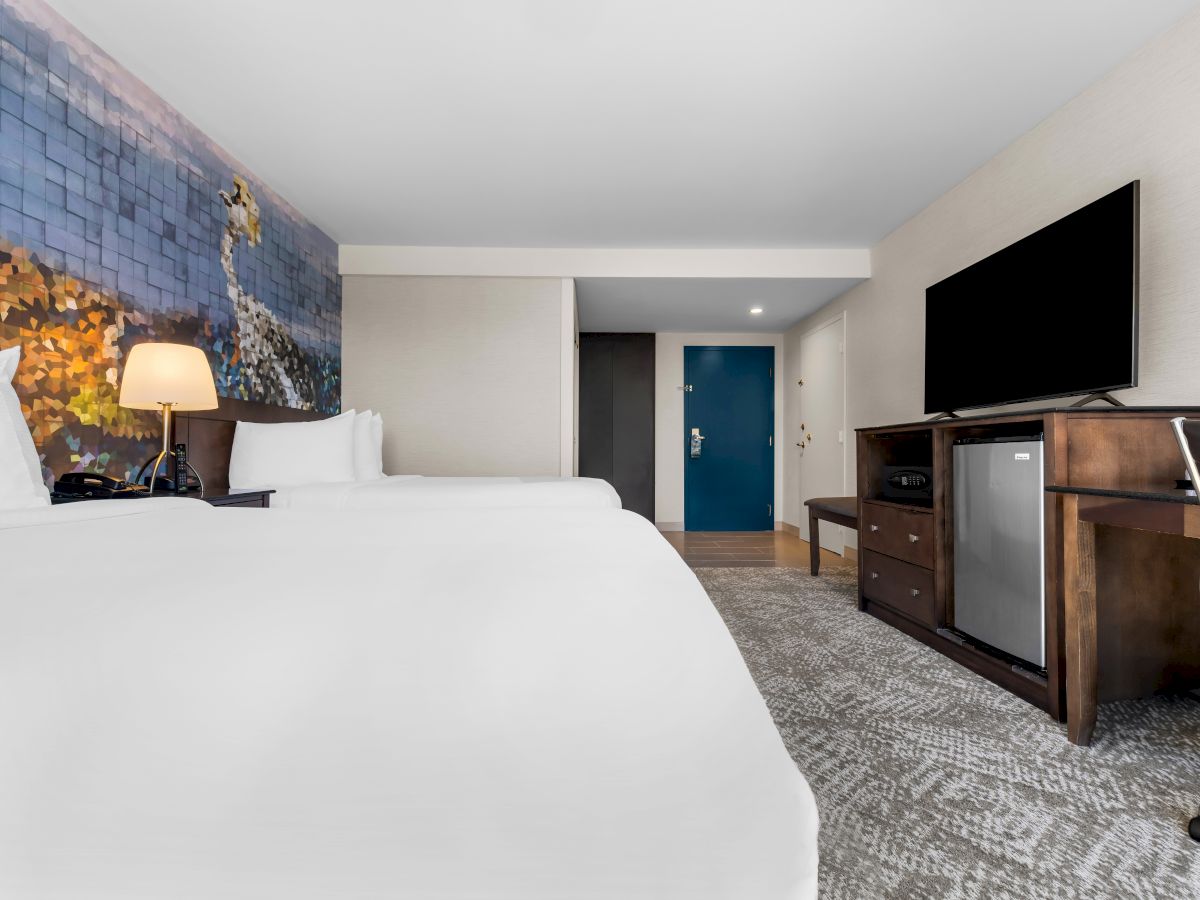 The image shows a modern hotel room with two beds, a mural, a TV, a mini fridge, and a desk with a chair, creating a cozy atmosphere.