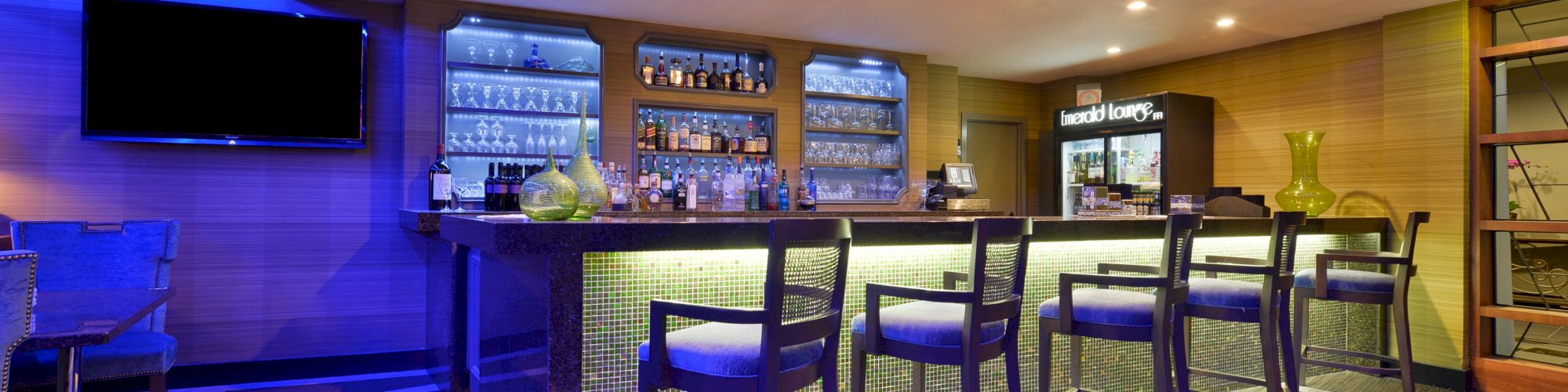 A modern bar with blue lighting, high stools, a TV, and a well-stocked counter, located in a sophisticated setting.