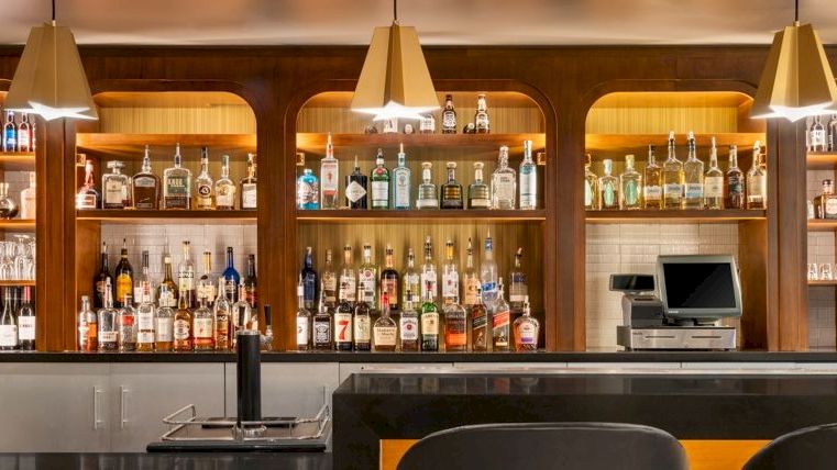A stylish bar with shelves of liquor bottles, pendant lights, and a counter with two TVs behind it.