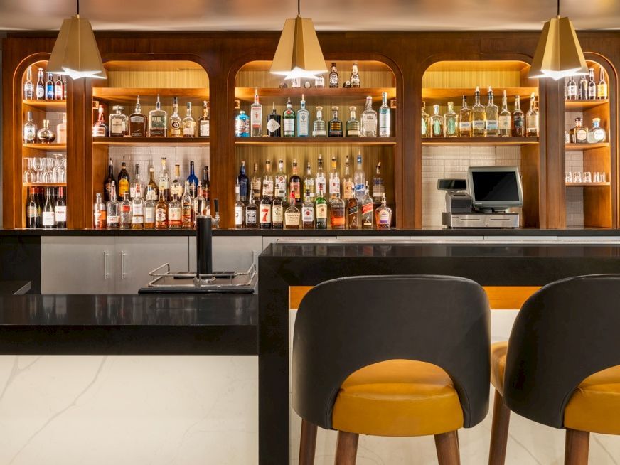 A hotel bar with shelves of liquor bottles, sleek countertops, pendant lights, and two yellow-cushioned stools.