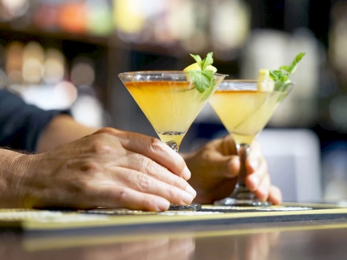 Two hands holding martini glasses filled with cocktails, garnished with lime and mint, in a dimly lit bar setting.