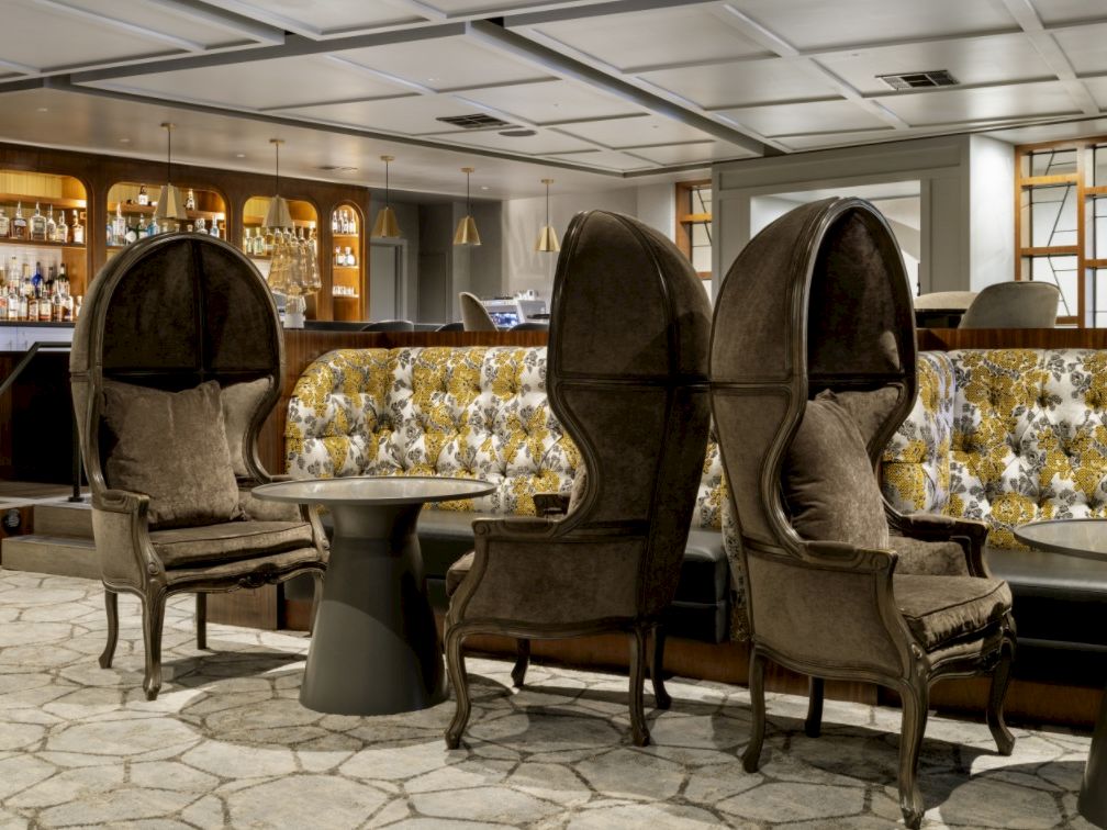 A hotel bar lounge with velvet tufted armchairs and patterned banquettes in front of a stocked liquor shelf.