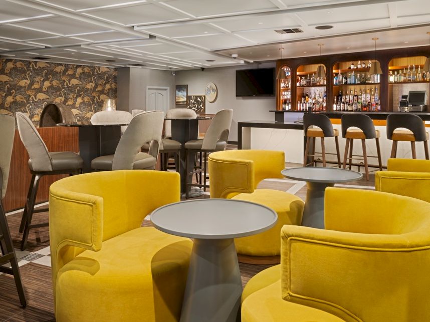 A modern bar lounge with yellow velvet seating, round tables, and a back counter stocked with bottles.