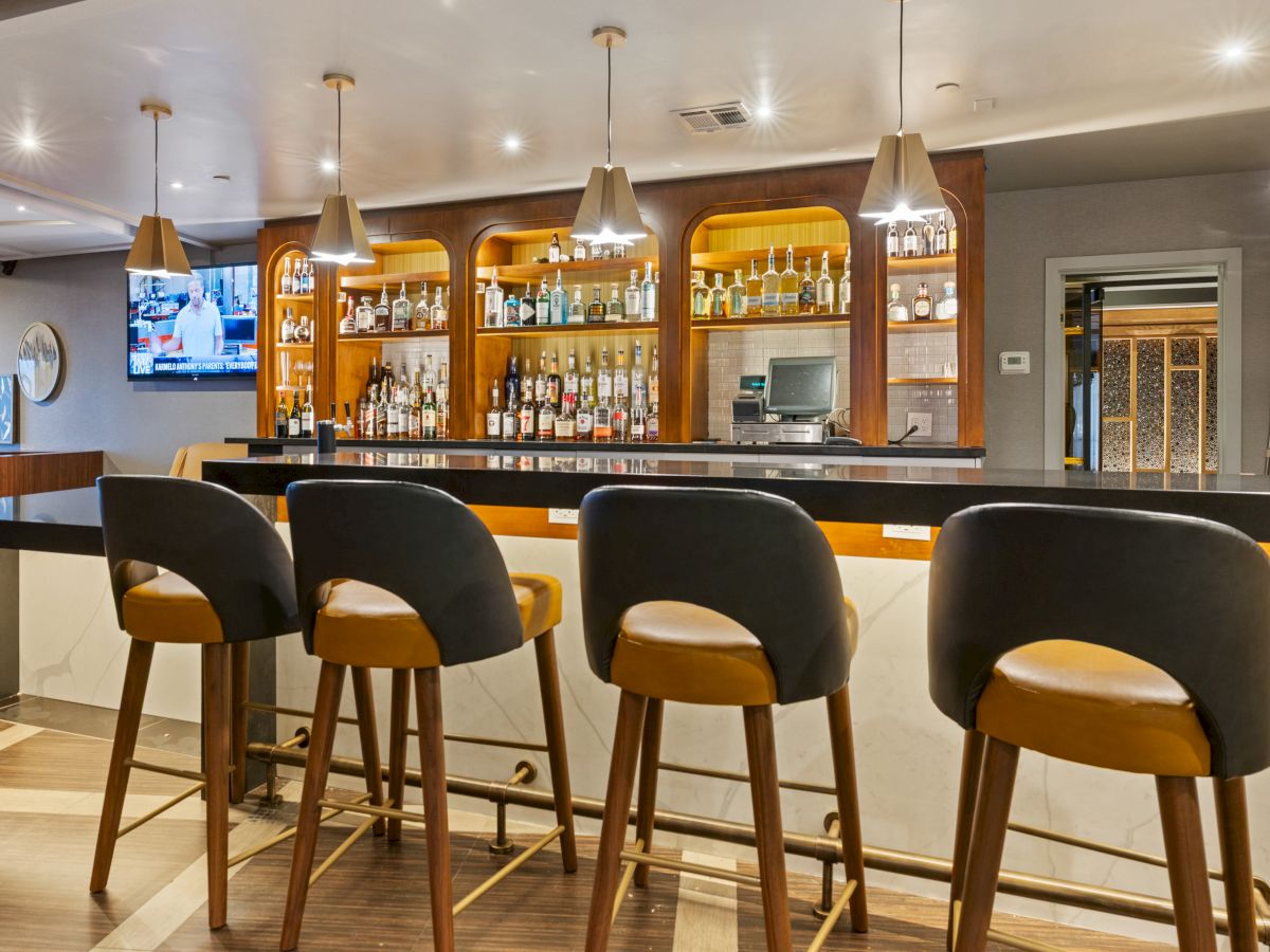 A modern bar with lit shelves of liquor behind a counter, pendant lights, and stools.