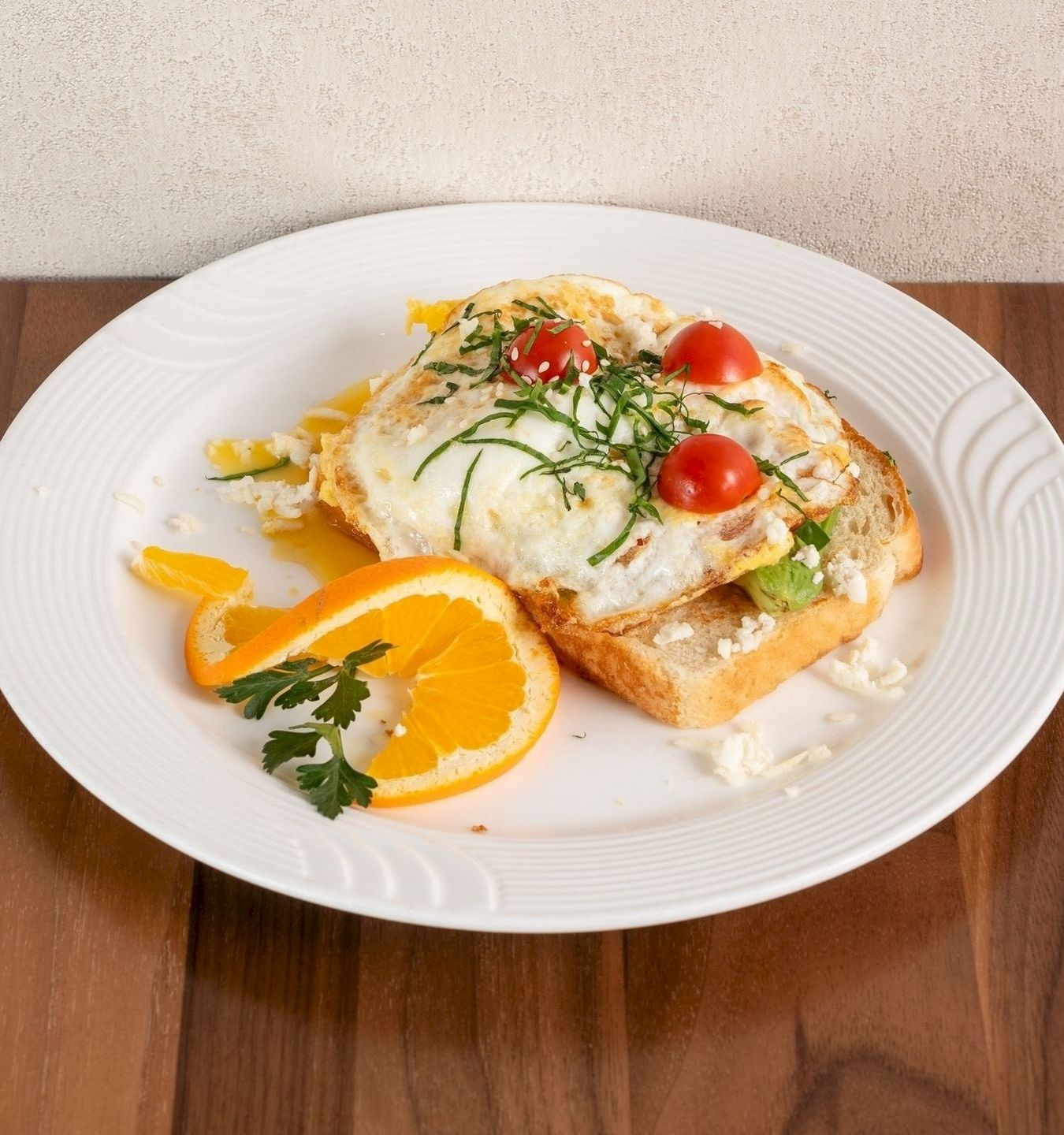 A white plate with two sunny-side eggs on toast, topped with tomatoes, sprouts, herbs, and orange slices.
