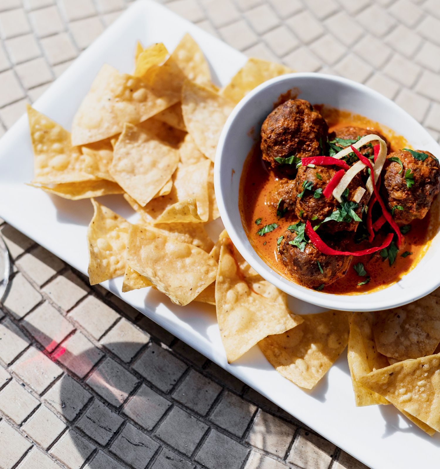 A bowl of meatballs in sauce sits on a plate with tortilla chips, accompanied by a glass of red wine on a tiled surface.