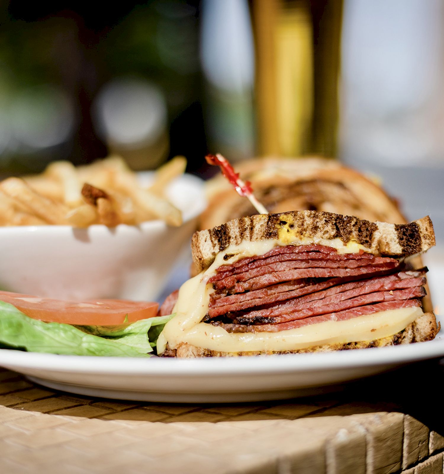 The image shows a sandwich with layers of meat and cheese, served with a side of fries in a bowl, and garnished with lettuce and tomato on a plate.