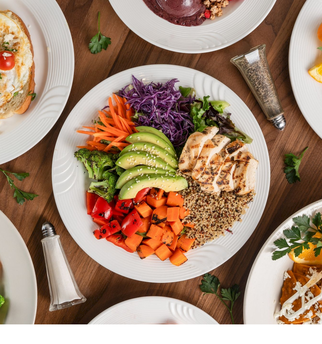 A spread of assorted meals&mdash;salad bowls, grilled chicken and roasted veggies, toast, dessert and fruit&mdash;on white plates.