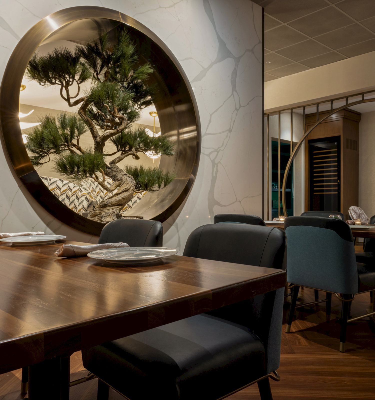 Modern dining room with dark chairs and a glowing bonsai tree displayed in a circular wall window.