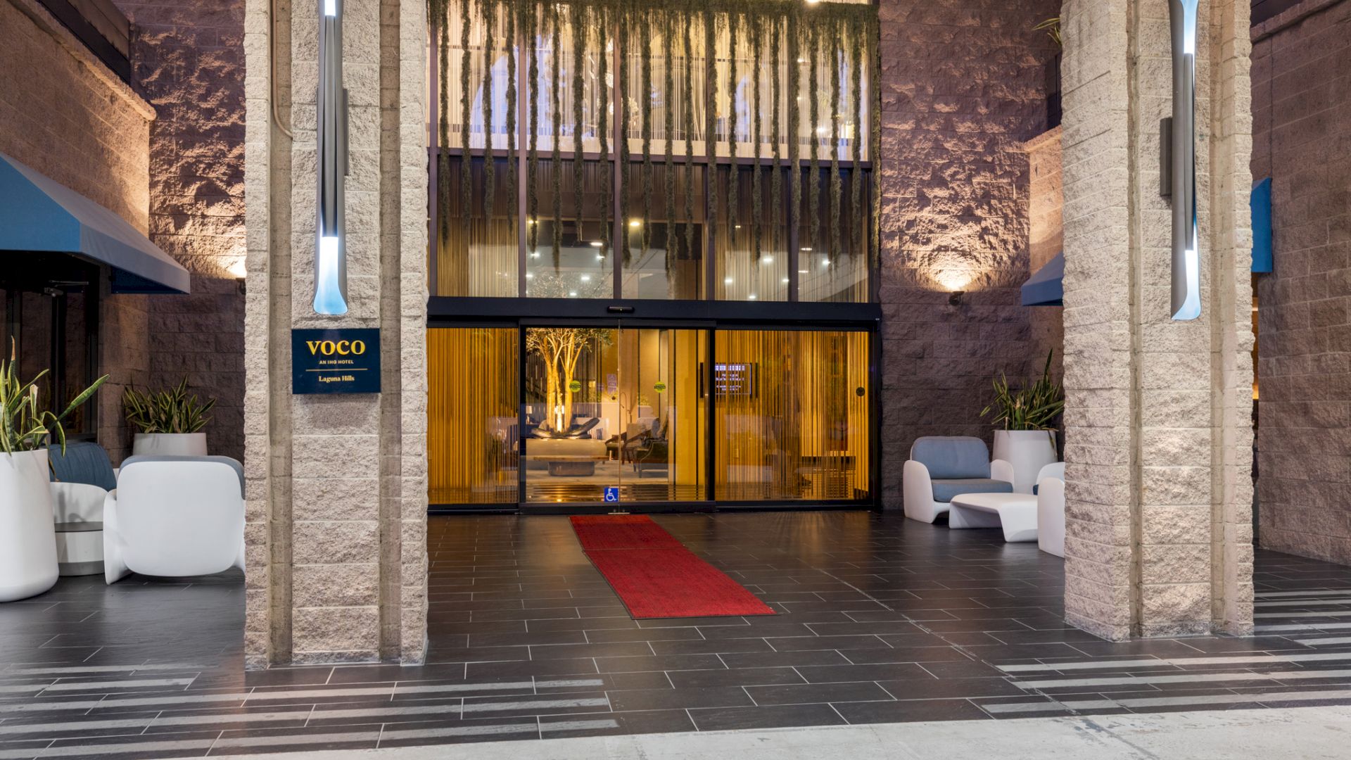 VOCO Laguna Hills hotel entrance with glass doors, warm lighting, lush hanging greenery, and vertical illuminated signs flanking the entry.
