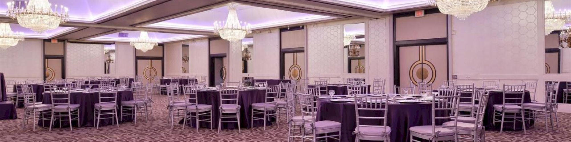 A large, elegant banquet hall with chandeliers, purple-lit ceiling, patterned carpet, and round tables covered with purple tablecloths surrounded by chairs.