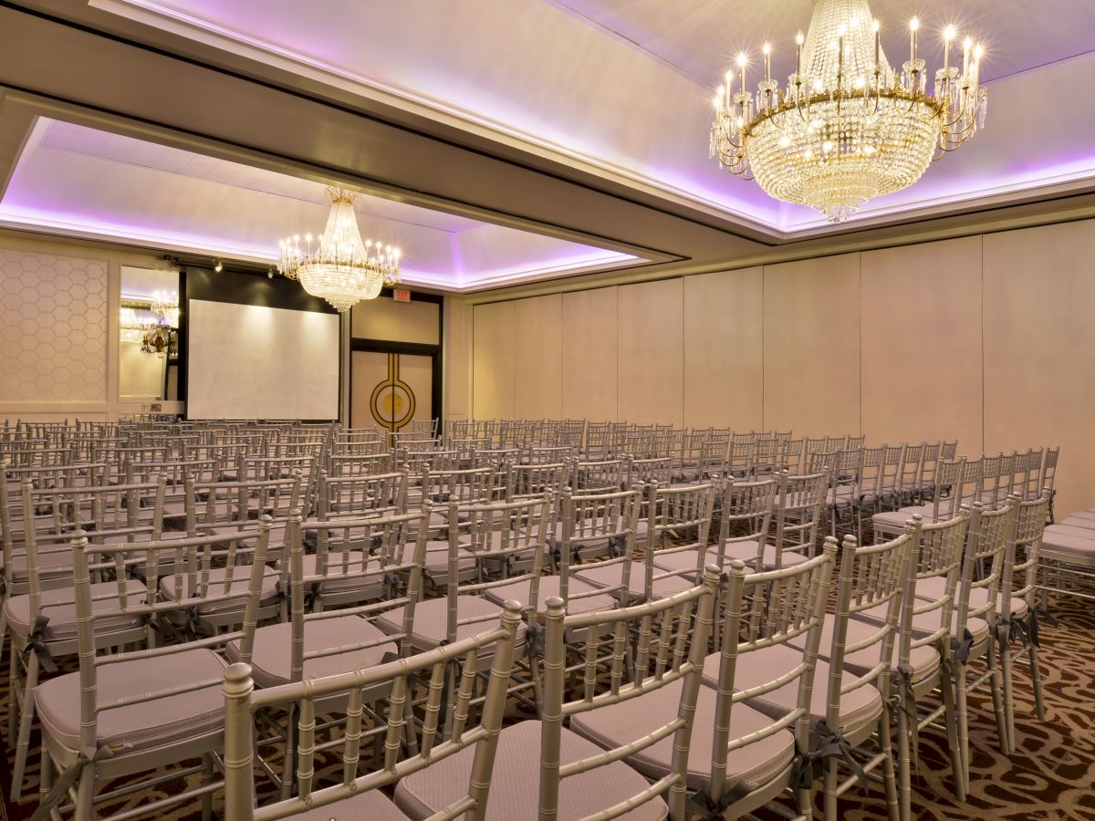 An elegant conference room with multiple rows of white chairs, chandeliers, a projector screen, and purple ambient lighting is prepared for an event.