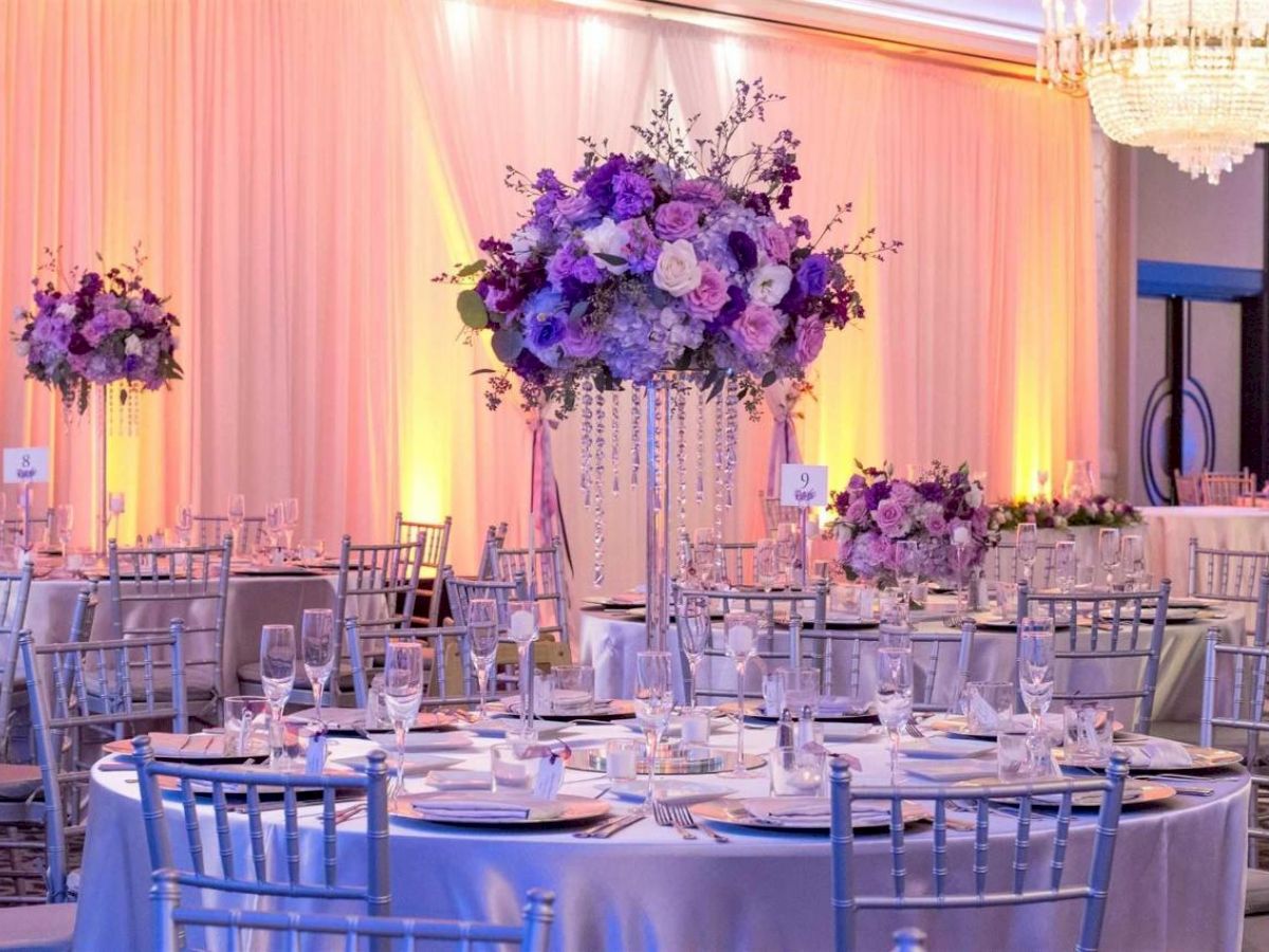 A decorated event hall with elegant round tables, purple floral centerpieces, silver chairs, soft lighting, and a chandelier.