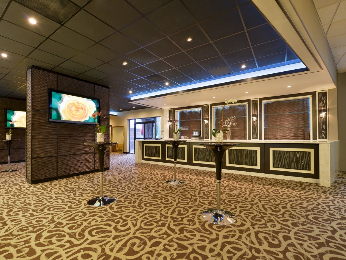 The image shows a modern hotel lobby with a patterned carpet, a reception desk, stools, and large TV screens displaying images.