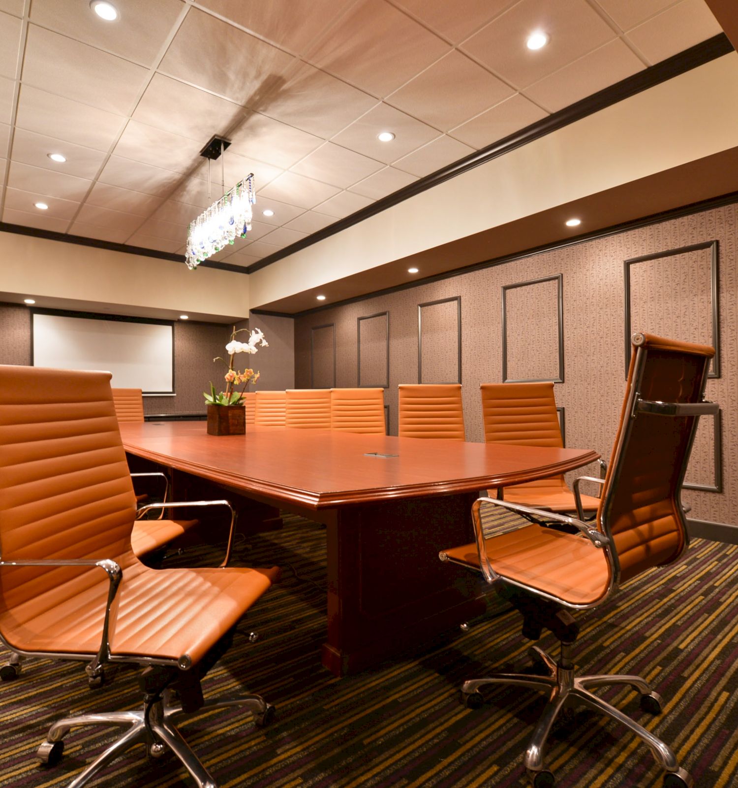 A modern conference room with a large wooden table, orange executive chairs, a whiteboard, a projector, and ceiling lights.