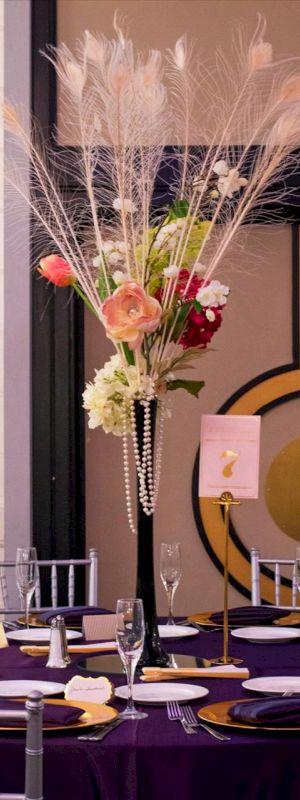 The image shows an elegantly set table with a tall floral centerpiece, wine glasses, and a table number holder. The decor is sophisticated and stylish.