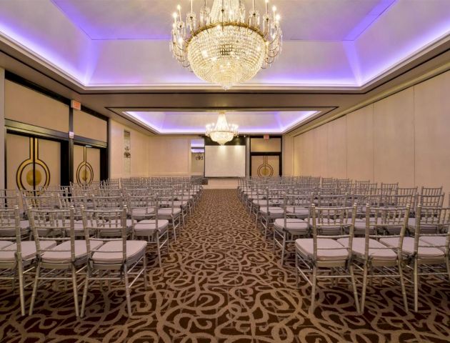 The image shows a conference room or ballroom set up with rows of chairs, a large chandelier, and a screen at the front, under purple lighting.