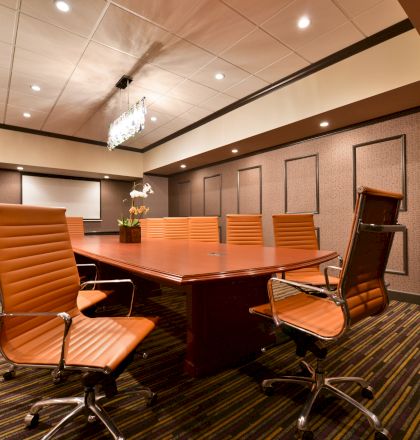 A modern conference room with a rectangular table, leather office chairs, a projector screen, and a small centerpiece on the table.