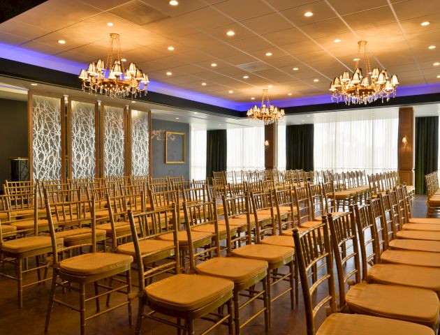 The image shows an elegantly decorated room with rows of empty chairs, chandeliers, and soft lighting, likely set up for an event or ceremony.