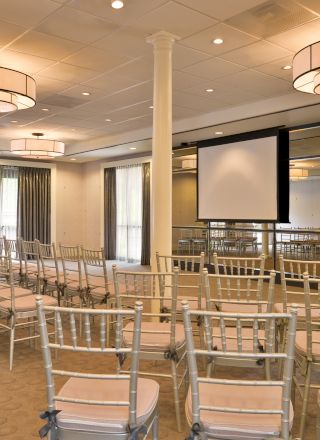 A conference room with rows of chairs facing a projection screen, mirrors on the walls, and elegant lighting fixtures hanging from the ceiling.