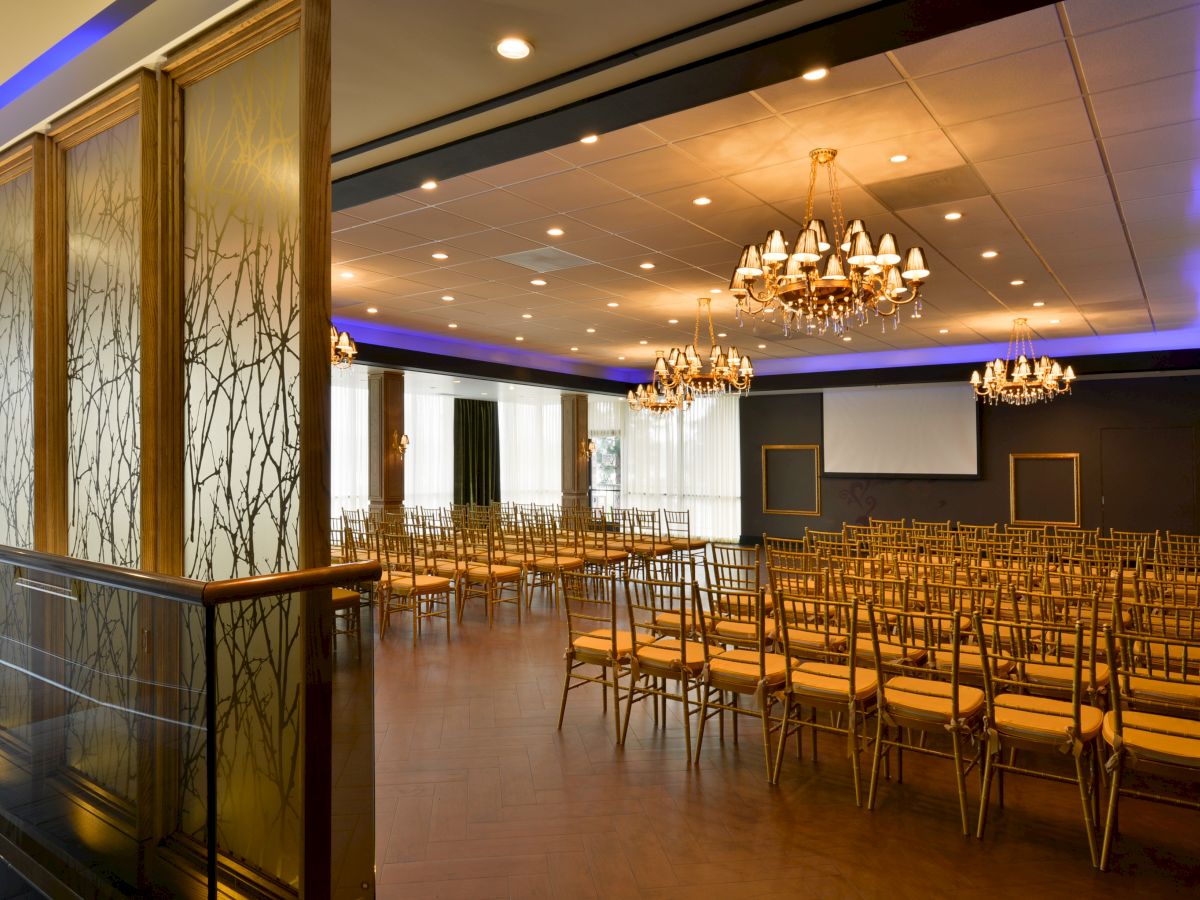 The image shows a well-lit, elegant event hall set with rows of chairs, chandeliers on the ceiling, and a screen at the front.