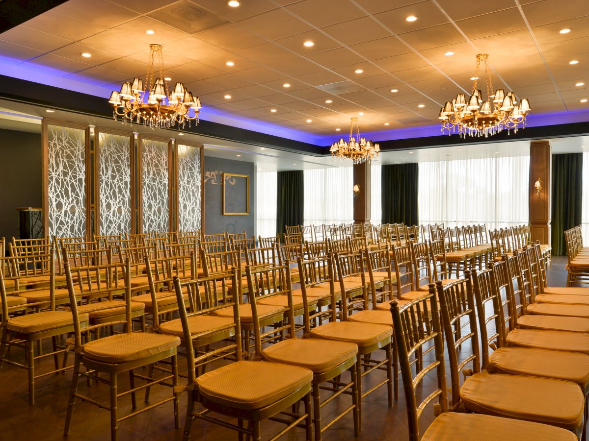 The image shows an elegant, empty event hall with rows of gold chairs, chandeliers, and a partition with an intricate design, under warm lighting.