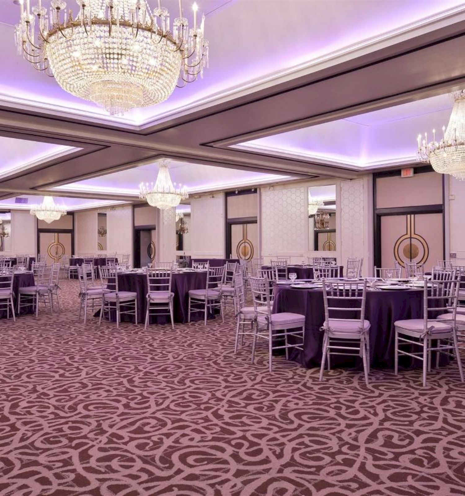 An elegant banquet hall with chandeliers, round tables covered in purple tablecloths, and silver chairs set up for an event, creating a luxurious ambiance.